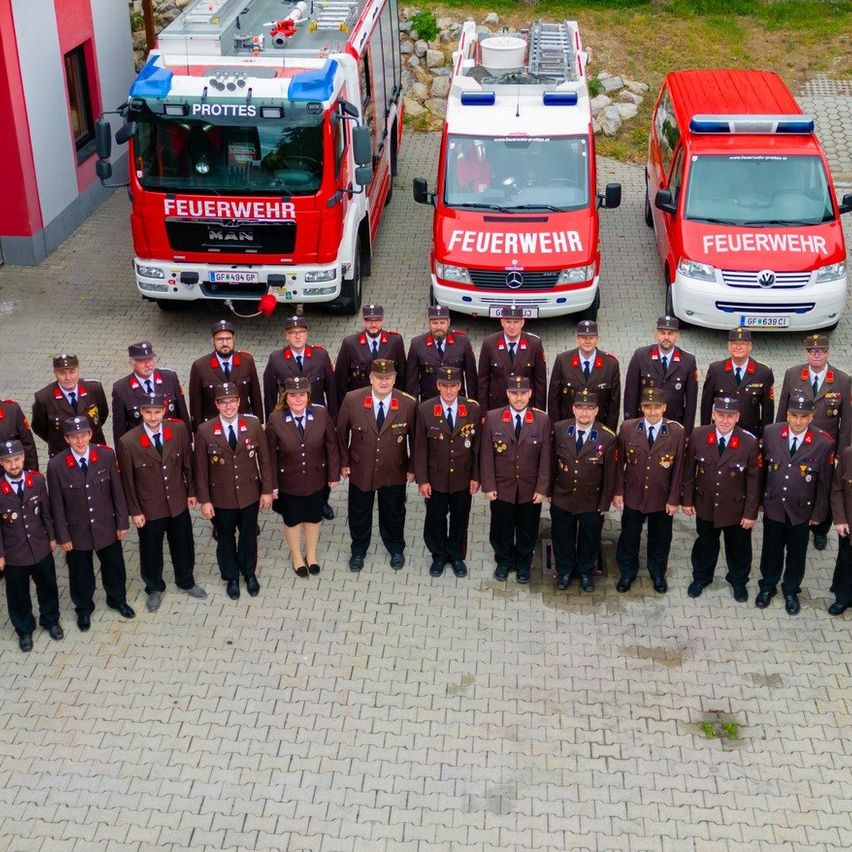 Eine Gruppe Feuerwehrleute in Uniform posiert für ein Foto vor ihren Feuerwehrfahrzeugen. Die Szene spielt sich in einem ländlichen Bereich ab, mit Gebäuden im Hintergrund.