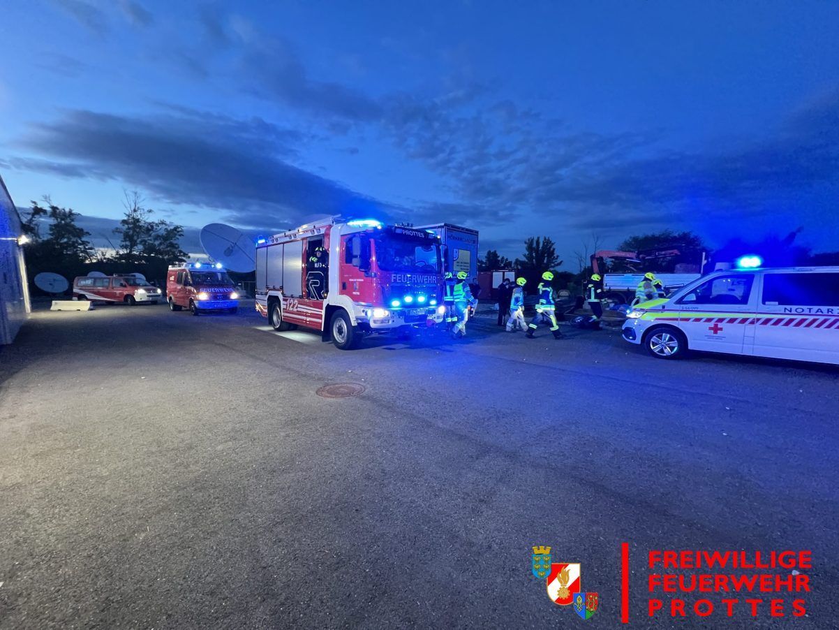 Rettungsfahrzeuge, einschließlich eines Feuerwehrwagens, sind auf einer Straße bei Dämmerung geparkt. Mehrere Personen in Schutzausrüstung kümmern sich um eine Situation. Ein Wasserzeichen mit dem Text 'FREIWILLE FEUERWEHR PROT' befindet sich in der unteren rechten Ecke.