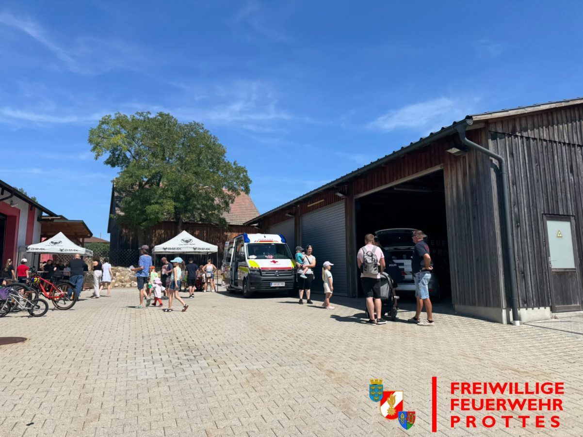 Eine Gruppe von Menschen versammelt sich vor einer Feuerwache, mit einem Krankenwagen in der Nähe. Einige stehen in der Nähe eines Zeltes, während andere in der Nähe der Garage sind. Der Himmel ist blau mit ein paar Wolken.