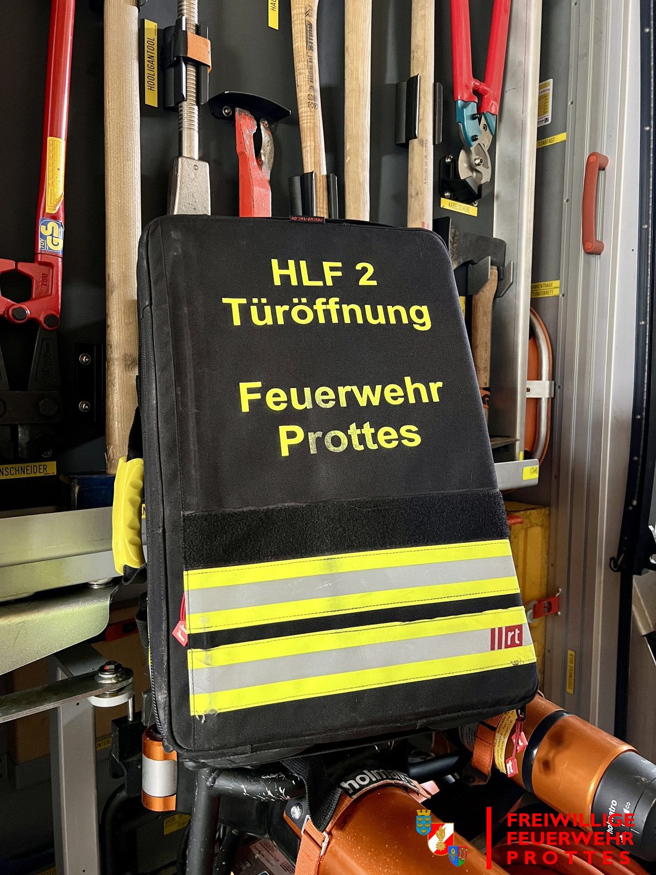 Ein schwarzer Beutel mit gelben und grauen Streifen, beschriftet mit 'HLF 2 Turoffnung Feuerwehr Protes', hängt an einem Metallregal in einem Lagerraum.