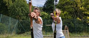 Ein Mann und eine Frau halten Bögen in einem Grasfeld mit einem Zaun und Bäumen im Hintergrund. Der Mann trägt ein weißes ärmelloses Hemd und graue Hose, während die Frau ein weißes ärmelloses Hemd und blaue Jeans trägt.