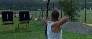 Eine Person übt Bogenschießen auf einer Wiese, mit zwei aufgestellten Holzzielen und Bergen im Hintergrund, während der Himmel von Orange zu Blau übergeht.