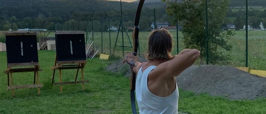 Eine Person übt Bogenschießen auf einer Wiese, mit zwei aufgestellten Holzzielen und Bergen im Hintergrund, während der Himmel von Orange zu Blau übergeht.