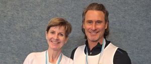 Ein Mann und eine Frau stehen zusammen und tragen beide weiße Hemden mit Medaillen um den Hals. Die Frau hat kurze Haare und lächelt, während der Mann eine längere Frisur hat und ebenfalls lächelt. Sie stehen vor einer grauen Wand.