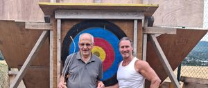 Two men stand in front of a makeshift archery target, one holding a bow and the other wearing a white tank top.