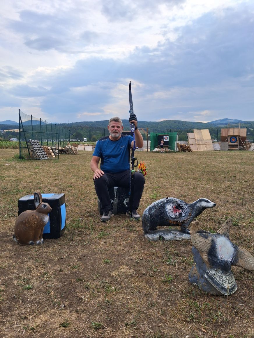 Ein Mann in einem blauen Shirt und schwarzen Hosen sitzt auf einer Bank in einem Außenbereich und hält einen Bogen und einen Pfeil. Vor ihm stehen drei Tierstatuen und auf der linken Seite befindet sich eine Aufbewahrungsbox. In der Ferne gibt es einen Zaun, ein Ziel und ein Gebäude.