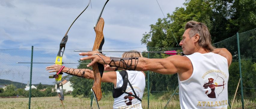 Zwei Personen, ein Mann und eine Frau, üben Bogenschießen im Freien. Sie stehen auf einem Grasfeld mit einem Zaun im Hintergrund. Beide tragen ärmellose Shirts mit Bogenschießmotiven.