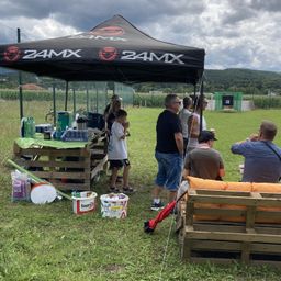 Eine Gruppe von Menschen steht unter einem 24MX-Zelt auf einem Rasenfeld. Ein Tisch mit verschiedenen Gegenständen ist in der Nähe aufgestellt. Der Hintergrund zeigt einen eingezäunten Bereich mit einem Schießstand.