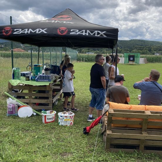 Eine Gruppe von Menschen steht unter einem 24MX-Zelt auf einem Rasenfeld. Ein Tisch mit verschiedenen Gegenständen ist in der Nähe aufgestellt. Der Hintergrund zeigt einen eingezäunten Bereich mit einem Schießstand.