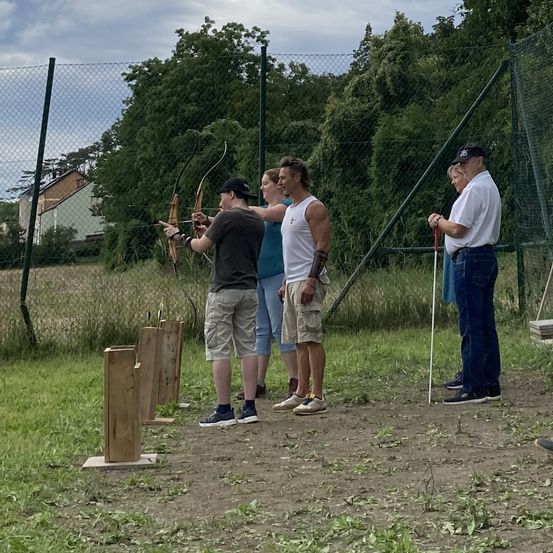 Eine Gruppe von Menschen übt Bogenschießen im Freien. Zwei Männer zielen mit ihren Bögen, während zwei andere zusehen. Eine Frau hält einen Stock. Dahinter steht ein Zaun und ein Feld.
