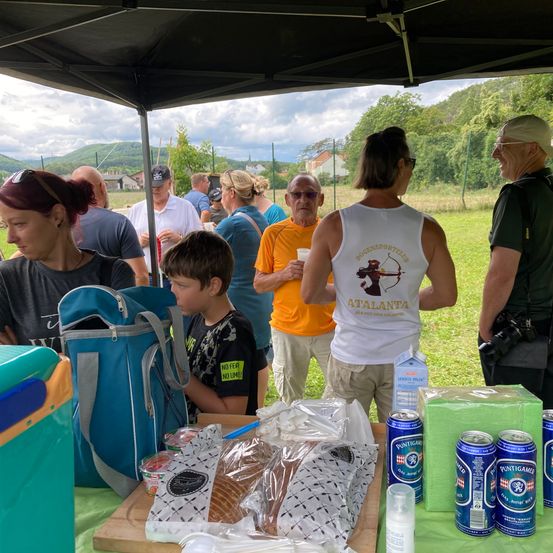 Eine Gruppe von Menschen versammelt sich unter einem Zelt auf einer Wiese. Eine Frau steht in der Nähe eines Tisches mit Essen, und ein Mann mit einer Kamera ist in der Nähe. Ein Junge schaut auf den Tisch und ein Mann im orangefarbenen Hemd. Im Hintergrund sind weitere Menschen zu sehen, und ein Berg ist sichtbar.