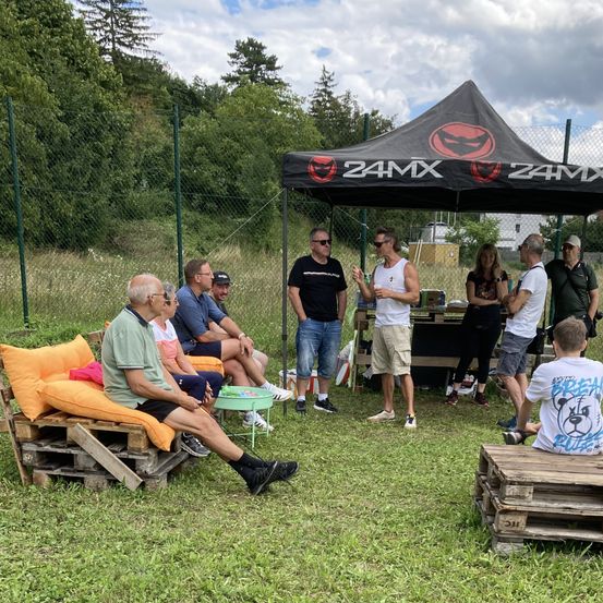 Mehrere Männer und Frauen versammeln sich unter einem Zelt mit dem 24MX-Logo, plaudern und sitzen auf Kissen. Ein Tisch steht in der Nähe mit verschiedenen Gegenständen.