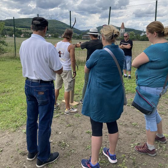 Eine Gruppe von Menschen steht in einem Feld mit einem Zaun, wahrscheinlich mit Bogenschießen beschäftigt. Eine Person zielt mit einem Bogen, während eine andere zuhört. Ein Grasbereich, ein Gebäude und ein Berg sind im Hintergrund.