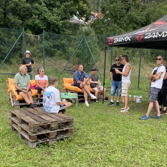 Eine Gruppe von Menschen versammelt sich unter einem Zelt in einem Park. Einige sitzen auf Paletten, während andere herumstehen. Sie sind von Bäumen und einem Zaun umgeben.