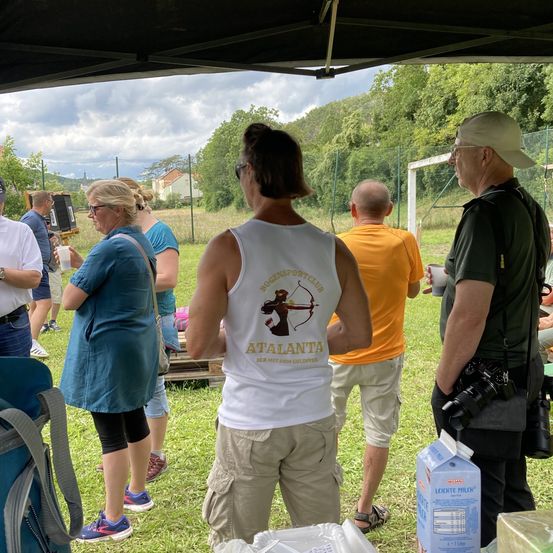 Mehrere Personen versammeln sich unter einem Zelt. Eine Frau in einem ärmellosen Shirt spricht mit einem Mann mit einer Kamera. Dahinter stehen andere in einem Grasfeld mit einem Fußballtor und Bäumen in der Ferne.