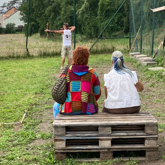 Zwei Frauen sitzen auf einem Holzpalette und beobachten einen Mann, der einen Bogen und Pfeile in einem Feld hält. Dahinter befindet sich ein Zaun und einige Werkzeuge.