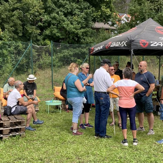Eine Gruppe von Menschen versammelt sich in einem Außenbereich unter einem Zelt, einige sitzen und andere stehen und unterhalten sich.