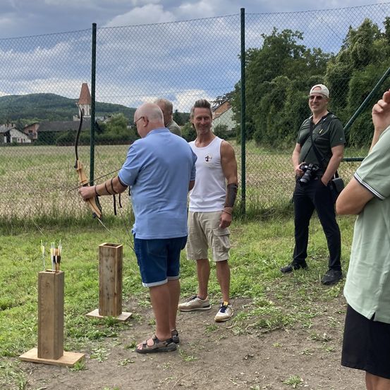 Fünf Männer stehen in einem Feld mit einem Zaun hinter ihnen. Ein Mann hält einen Bogen und eine Pfeil. Ein anderer Mann macht ein Foto mit einer Kamera.