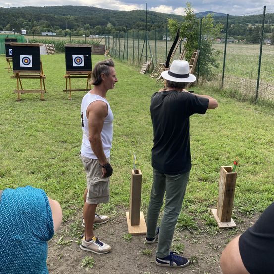 Zwei Männer sind an einer Bogenschießaktivität im Freien beteiligt. Ein Mann bereitet sich auf den Schuss vor, während der andere zusieht. Hinter ihnen stehen Holzpfeile auf einem Rasenfeld.