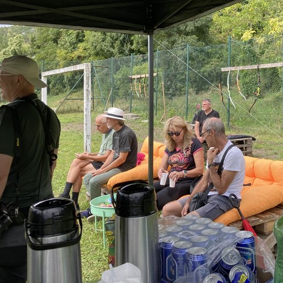 Eine Gruppe von Menschen sitzt auf einer orangefarbenen Couch unter einem Zelt, wahrscheinlich bei einem geselligem Treffen. Ein Mann steht in der Nähe mit einer Kamera. In der Nähe gibt es Becher, einen Tisch und eine Flasche. Im Hintergrund befinden sich einige Bäume, ein Zaun und ein Bogenschießziel.
