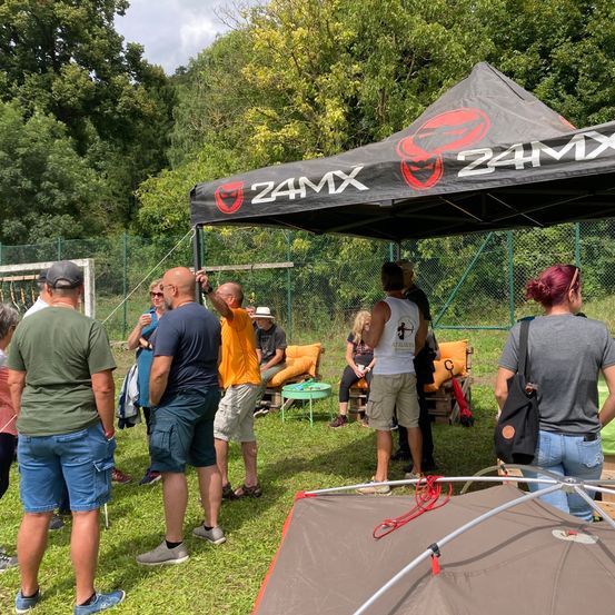 Eine Gruppe von Menschen versammelt sich unter einem 24MX-Zelt, wahrscheinlich für eine Veranstaltung. Einige stehen, andere sitzen auf Stühlen. Eine Frau mit einer Tasche ist dabei. Das Gebiet ist von einem Zaun umgeben, dahinter befinden sich Bäume und ein Grasfeld.