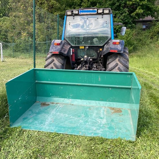 Ein blauer Traktor mit einer grünen Kiste steht auf einem Rasenfeld, umgeben von einem Zaun. Hinter dem Zaun befinden sich Bäume und ein Haus.