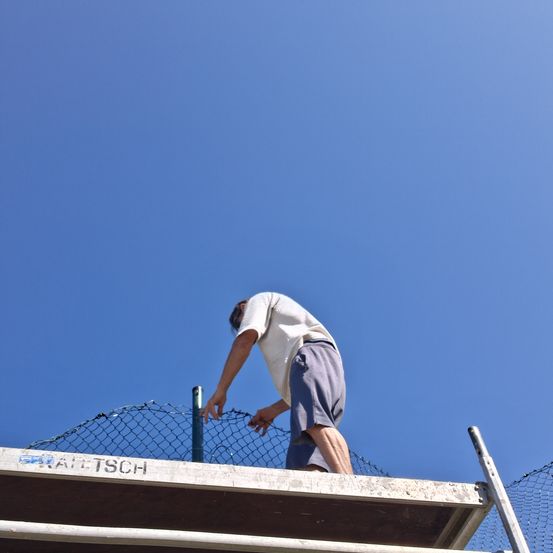 Ein Mann ist auf einem Dach und repariert ein Netzzaun unter einem klaren blauen Himmel.
