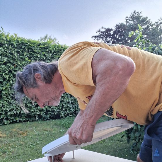 Ein älterer Mann in einem gelben T-Shirt und blauen Shorts beugt sich über ein weißes Objekt auf einem Holztisch, scheinbar wird es repariert. Die Szene spielt sich in einem Garten mit einer Hecke und Rasen ab.