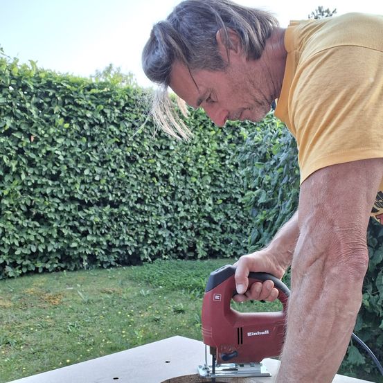 Ein Mann in einem gelben T-Shirt benutzt eine Stichsäge, um ein Holzbrett im Garten zu schneiden. Der Mann beugt sich über den Tisch und konzentriert sich auf seine Arbeit.