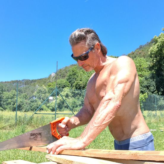 Ein oberkörperfreier Mann mit Sonnenbrille säg ein Stück Holz mit einer großen orangefarbenen Säge im Freien. Er steht auf Gras mit einem eingezäunten Bereich und Bäumen dahinter.