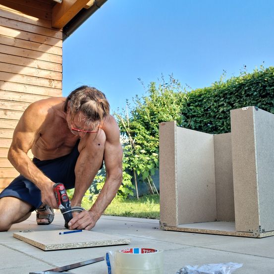 Ein Mann mit Brille und Shorts kniet auf dem Boden und benutzt einen Akkuschrauber, um an einer Holzplatte zu arbeiten. Ein großer Karton ist in der Nähe.