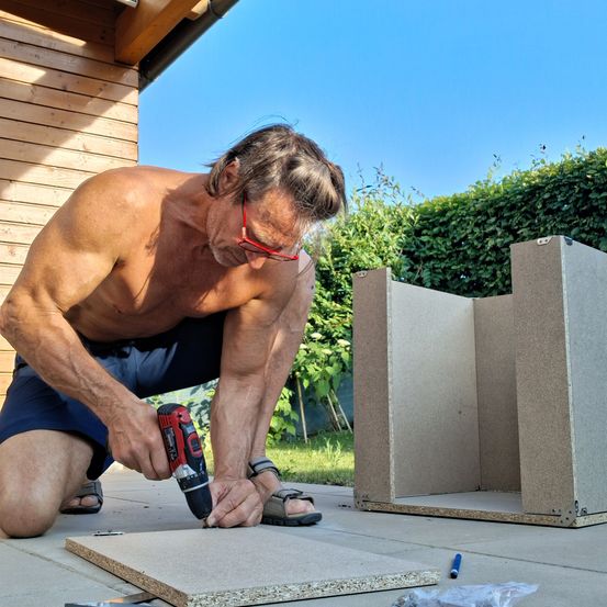 Ein Mann in Sonnenbrille und Shorts kniet und benutzt einen Akkuschrauber, um eine Holzplatte zu reparieren. Hinter ihm sind ein unfertiges Holzgerüst und eine Hecke bei klarem Himmel zu sehen.