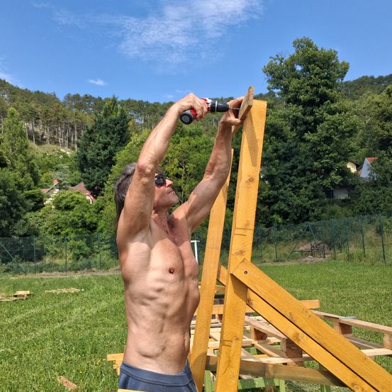 Ein Mann ohne Oberbekleidung benutzt einen Akkuschrauber, um eine Holzkonstruktion auf einer Wiese mit Bäumen und einem Hügel im Hintergrund zu montieren.