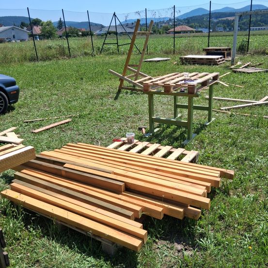 Auf einem Grasfeld sind mehrere Holzbretter gestapelt. Ein Holztisch mit einem Becher steht in der Nähe. Ein Holzstuhl ist hinter dem Tisch aufgestellt. Häuser und Berge sind in der Ferne zu sehen.