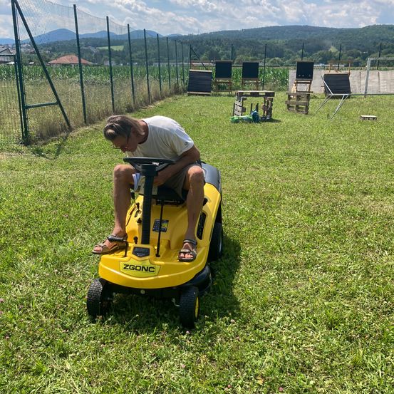 Ein Mann in Sandalen sitzt auf einem gelben Rasenmäher mit der Aufschrift 'ZGONC' vorne, wahrscheinlich in einem Grasbereich mit Zaun und Bergen im Hintergrund.