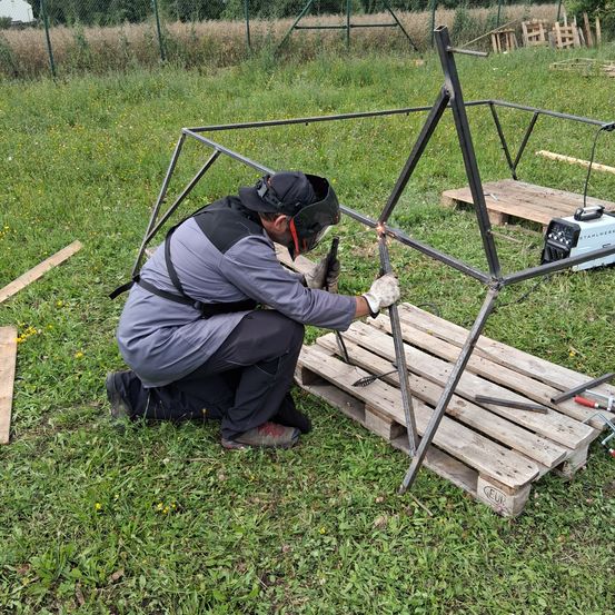 Ein Mann in einer Schweißmaske schweißt eine Metallstruktur auf einem Holzpalette in einem Grasfeld.