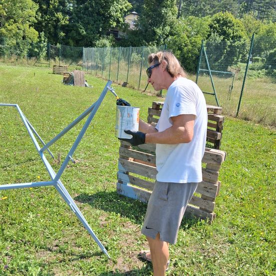 Ein Mann in Sonnenbrille und weißem T-Shirt malt einen Metallständer im Freien an einem sonnigen Tag.