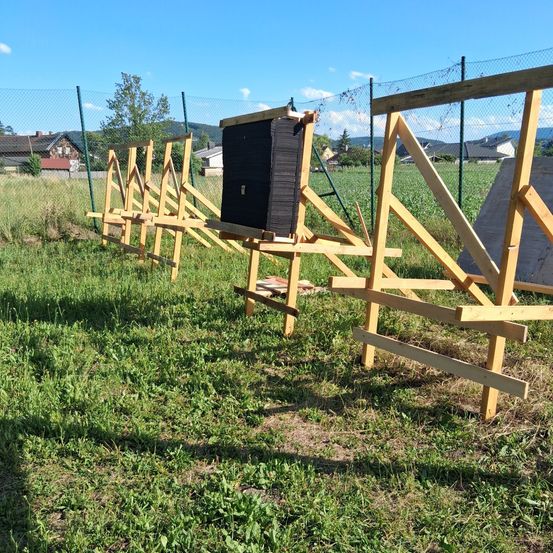 Eine Holzkonstruktion in einem Grasfeld mit einem Zaun und Häusern im Hintergrund. Der Himmel ist klar und blau.