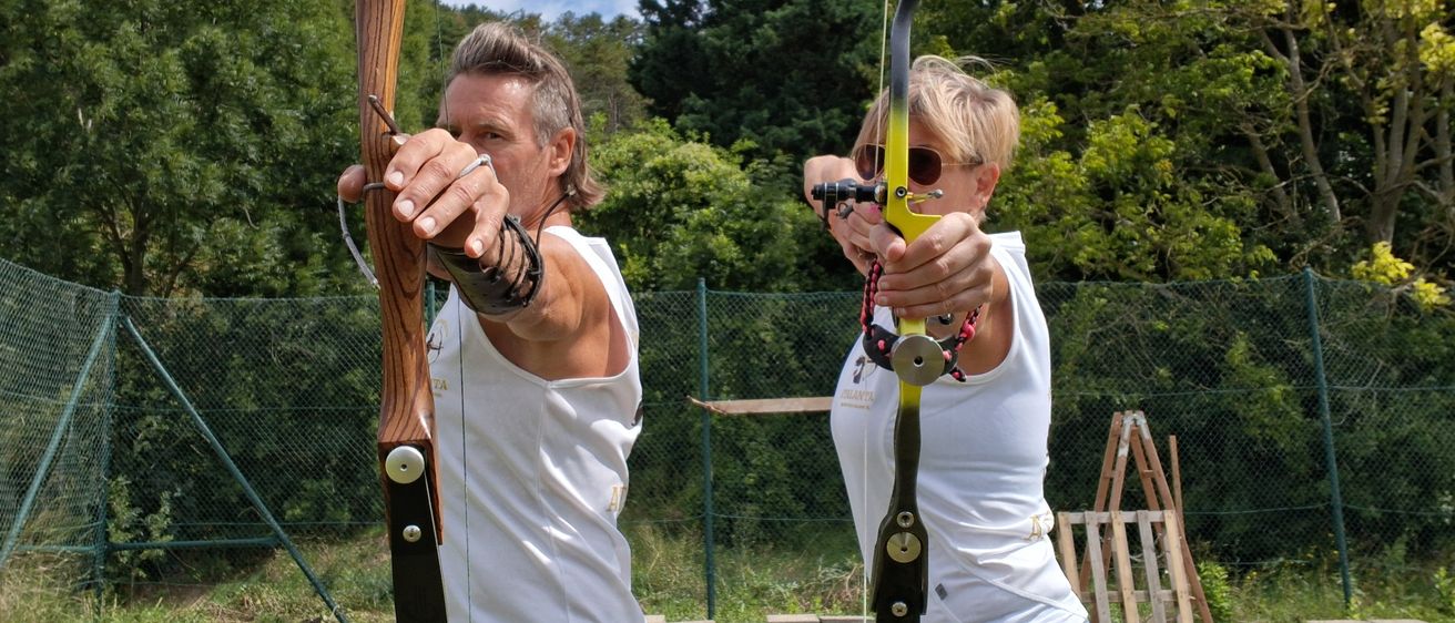 Zwei Personen, ein Mann und eine Frau, stehen auf einem Grasfeld, halten Bögen und posieren für ein Foto. Sie sind lässig gekleidet und scheinen an einer Bogenschießaktivität teilzunehmen.