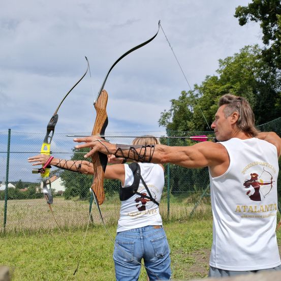 Zwei Personen, ein Mann und eine Frau, üben Bogenschießen auf einem Freiluftfeld. Sie halten beide Bögen und Pfeile. Der Mann trägt ein ärmelloses Shirt mit dem Wort Atalanta.