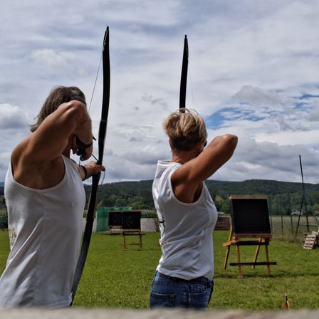 Zwei Personen zielen mit ihren Bögen in einer Freiluftumgebung, wahrscheinlich beim Bogenschießen. Ein grüner Hintergrund mit Zaun und Bergen ist hinter ihnen.