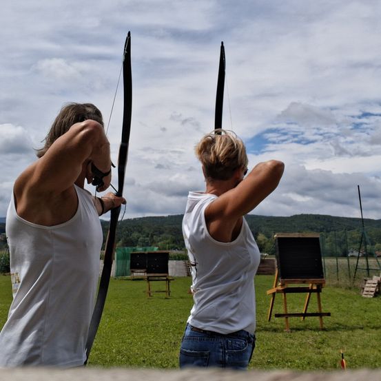 Zwei Personen zielen mit ihren Bögen in einer Freiluftumgebung, wahrscheinlich beim Bogenschießen. Ein grüner Hintergrund mit Zaun und Bergen ist hinter ihnen.