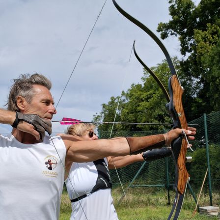 Ein Mann und eine Frau üben im Freien Bogenschießen und zielen mit einem rosa Pfeil auf ein Ziel. Der Mann trägt ein weißes ärmelloses Shirt mit Logo. Dahinter befindet sich ein Zaun und Bäume.