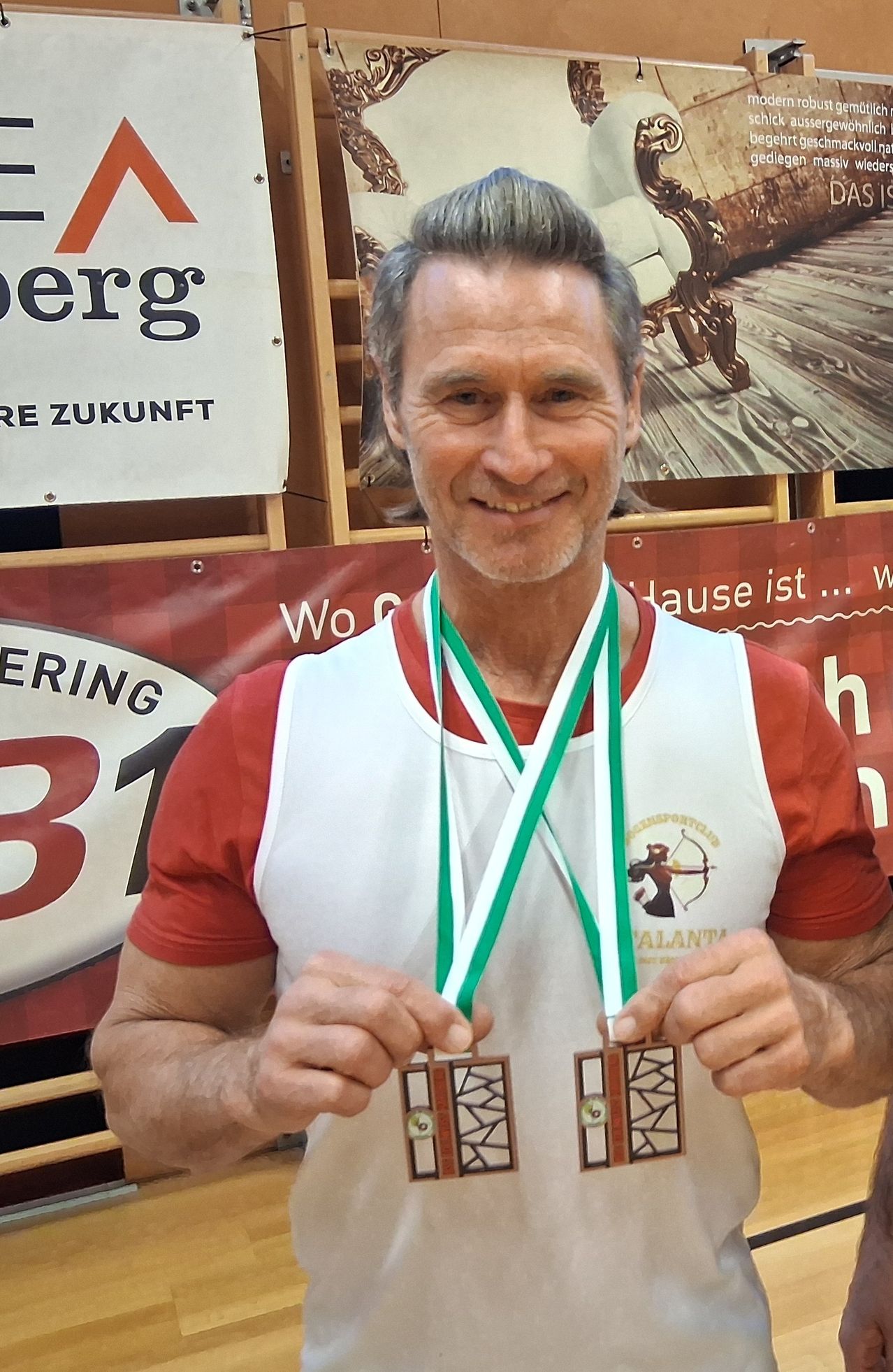 Ein Mann in einem roten Shirt und einer weißen Schürze hält zwei Medaillen in den Händen. Er lächelt und posiert für ein Foto. Hinter ihm befindet sich ein roter Banner mit der Nummer 31 und einigem Text.