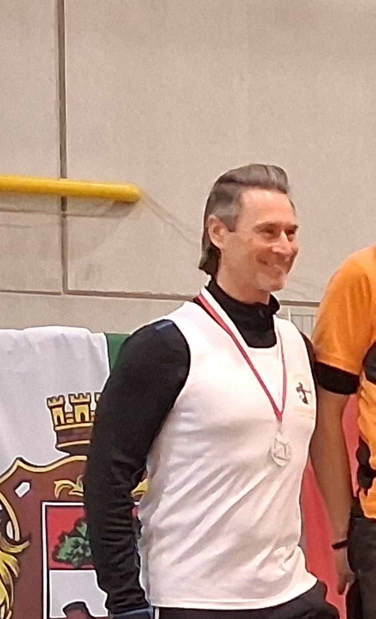 Ein Mann in einem weißen Shirt mit einer Medaille um den Hals steht neben einer anderen Person in einem orangefarbenen Shirt, beide lächeln. Der Hintergrund zeigt ein weißes Banner mit einem Logo und einem gelben Balken.