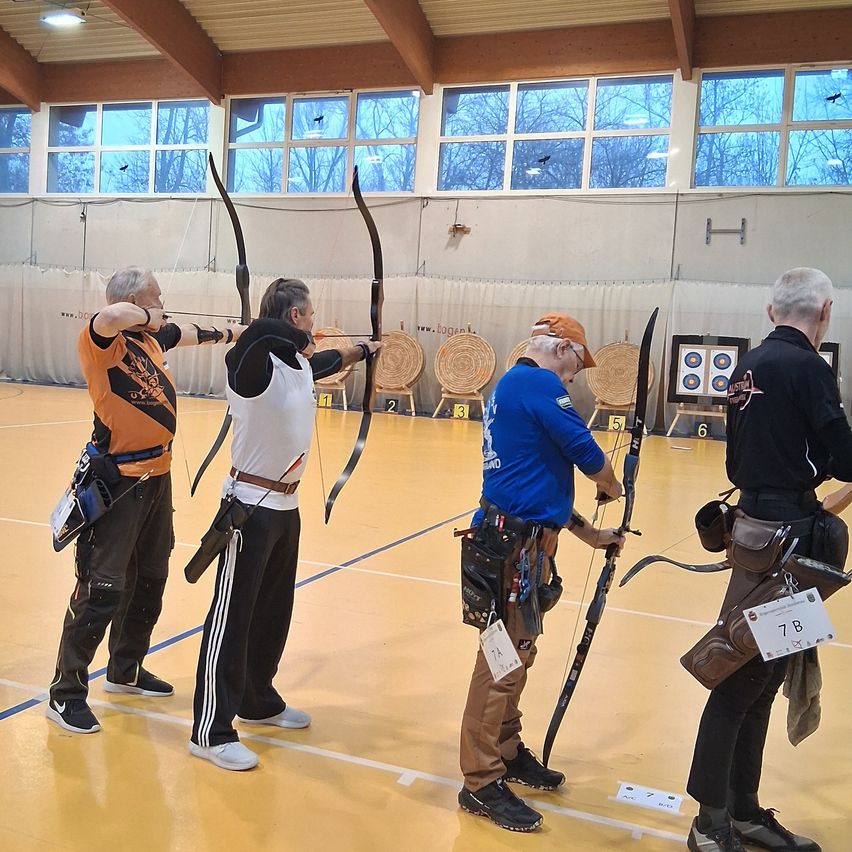 Vier Männer stehen in einer Turnhalle mit Bögen in der Hand und bereiten sich auf einen Bogenschießwettbewerb vor. Die Ziele sind im Hintergrund aufgestellt.