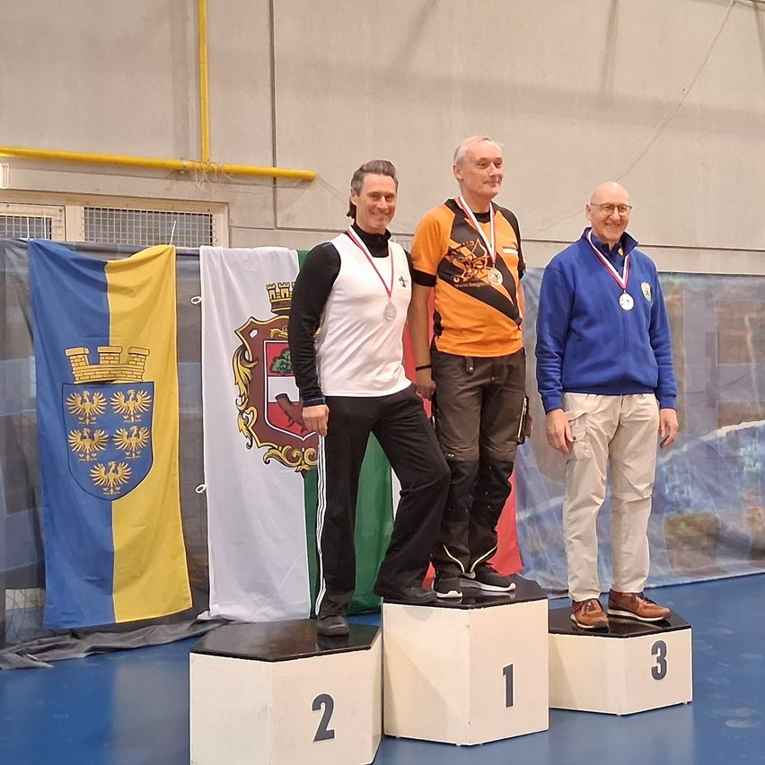Drei Männer stehen auf einem Podium mit nummerierten Stufen und tragen Medaillen. Der Mann auf der linken Seite trägt ein weißes Shirt und schwarze Hose, der Mann in der Mitte ein orangefarbenes Shirt und der Mann auf der rechten Seite einen blauen Pullover. Hinter ihnen befinden sich drei Flaggen, darunter eine mit Kronendesign.