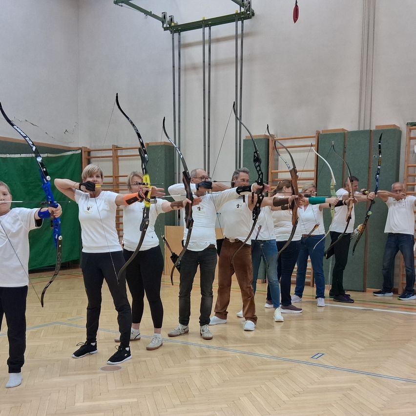Eine Gruppe von Menschen in weißen Shirts übt Bogenschießen in einer Turnhalle. Sie halten alle Bögen und Pfeile.