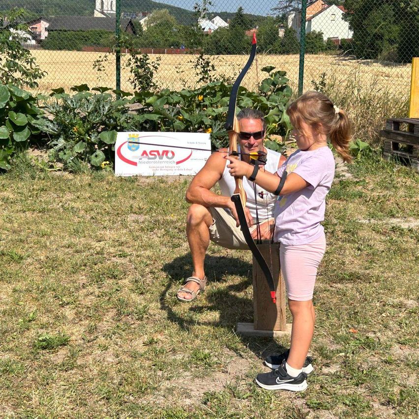 A man and a young girl are practicing archery outdoors. The man is kneeling and holding a bow while the girl stands beside him, holding her own bow.