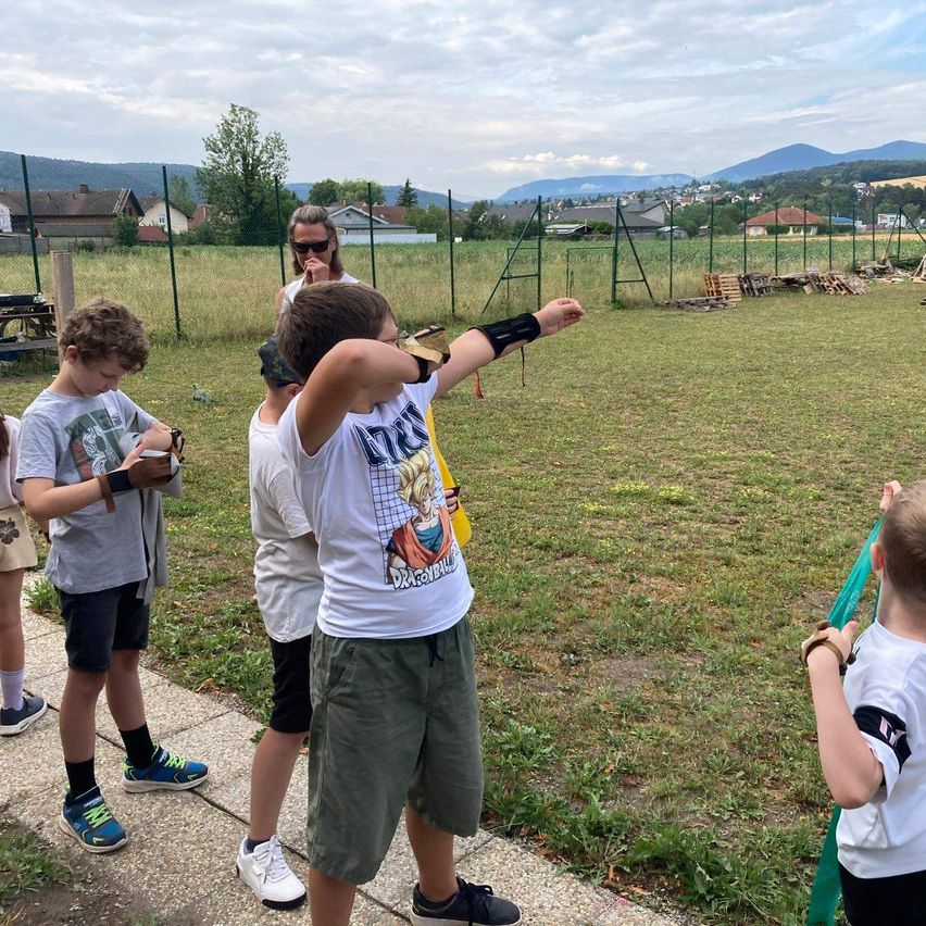 A group of children and an adult are standing in an open field, possibly practicing archery. The children are wearing protective gear.